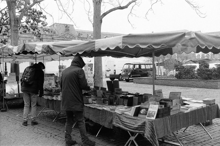 A street market stall with a large striped fabric table cover displaying an array of books arranged in stacks and rows under a canopy with scalloped edges. Two people are browsing the books; one person in a hooded jacket stands next to the stall with a backpack, while another in a coat appears to be examining the selections. The stall is situated on a cobblestone pavement beside a large tree, with additional market stalls visible in the background along with parked vehicles, including a small van, and a historic building with decorative elements and a colonnade. The scene is outdoors, during an overcast day, with leafless trees indicating a colder season. [COMPANY_NAME] may utilize similar logistical setups and staging, related to home relocation or furniture transport services, as seen in professional moving and packing scenarios.