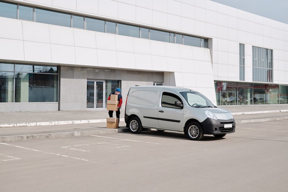 A person wearing a red jacket and blue beanie is seen unloading cardboard boxes from inside a grey delivery van parked in a commercial parking lot in front of a modern white building with large glass windows. The individual is carrying a box, with two additional boxes stacked on the pavement beside them. The van is positioned close to the building's entrance, with the rear open, suggesting a home relocation or furniture transport process. The parking lot is mostly empty, with painted lines for parking spaces, and the scene is set during daylight with overcast sky. This image illustrates a typical unloading stage during a house removals service in an urban area, facilitated by [COMPANY_NAME], focusing on packing and moving logistics.