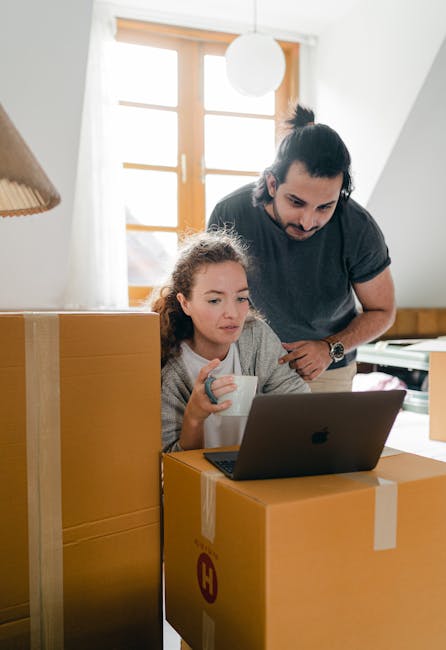 A man with dark hair tied back and wearing a dark grey T-shirt is supervising a home relocation process inside a bright room with wooden window frames and a white ceiling. He is leaning slightly forward with his hands on a large cardboard box, which is sealed with packing tape and positioned on the floor. Next to him, a young girl with curly light brown hair, dressed in a grey sweater, is sitting at a table or counter, holding a white mug in her left hand while looking at a laptop placed on the box. Surrounding them are additional cardboard boxes, some stacked and others placed on the floor, which are likely used for packing belongings during the furniture transport and packing process with a professional removals service. The environment features natural lighting coming through the windows, and lighting fixtures, such as a white pendant light and a wooden lampshade, are visible in the room. This scene illustrates the logistics of home removals, including packing, supervising, and preparing items for transportation, with the assistance of companies like Man With a Van Harringay.