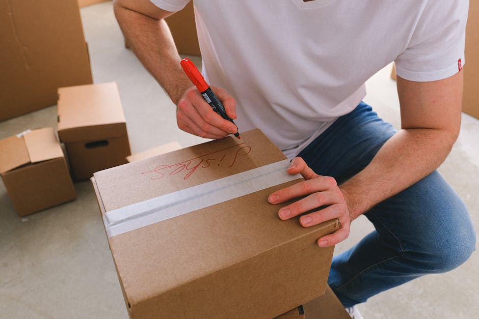A person wearing a white T-shirt and blue jeans is kneeling indoors beside a cardboard box, which is closed with clear packing tape across the top. The individual is using a red marker to write on the box's surface, with the word 'shoes' visible in red handwriting. In the background, several other cardboard boxes are stacked or placed on the floor, some of which are partially opened, revealing packing materials or contents inside. The setting appears to be a room prepared for house removal, with a clean, well-lit environment. The scene captures the packing and organization process involved in home relocation, suggesting careful handling of belongings before loading onto a vehicle. As part of [COMPANY_NAME]'s furniture transport and moving services, this illustrates the step of labeling boxes to ensure efficient unloading and placement during a professional move. The environment emphasizes a systematic packing approach typical of house removals, highlighting the importance of proper labeling and protective packaging materials for a smooth moving process.