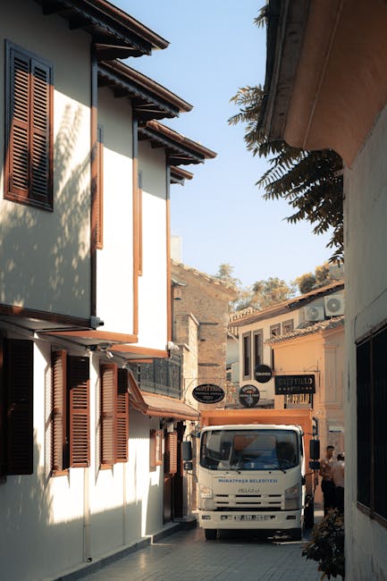 Photograph of a narrow residential street in Harringay during daytime, showing a white moving van parked between two closely spaced buildings with traditional wooden shuttered windows. The van is positioned with its front facing away from the viewer, and a person can be seen nearby, possibly involved in the home relocation process. The buildings feature light-colored facades, with some greenery visible over the roofs and along the street. The scene captures the challenge of maneuvering large furniture or packed boxes through tight access routes, emphasizing the importance of professional removals services from companies like Man With a Van Harringay for efficient furniture transport and packing and moving operations in confined spaces.