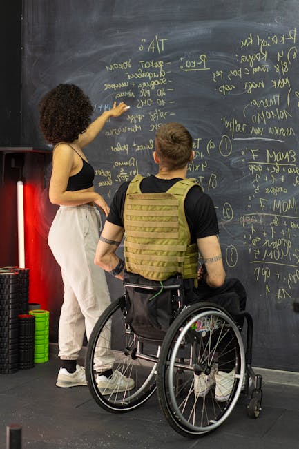 A woman with curly hair, wearing a black top and light-colored trousers, is standing and writing on a large blackboard filled with yellow chalk notes, diagrams, and calculations related to house removals and moving logistics. In front of her, a man in a black shirt with a tan tactical vest and seated in a wheelchair is observing her writing, with his hands resting on his lap. The scene appears to be inside a gym or training facility, with gym equipment, such as stacked weight plates and foam rollers, visible on the left side of the image. The environment is well-lit, and the focus is on the interaction between the woman and the man, reflecting discussions about packing, loading, and navigating narrow access during home relocation, a service provided by Man With a Van Harringay, aligning with the page about common problems with narrow access Harringay removals.
