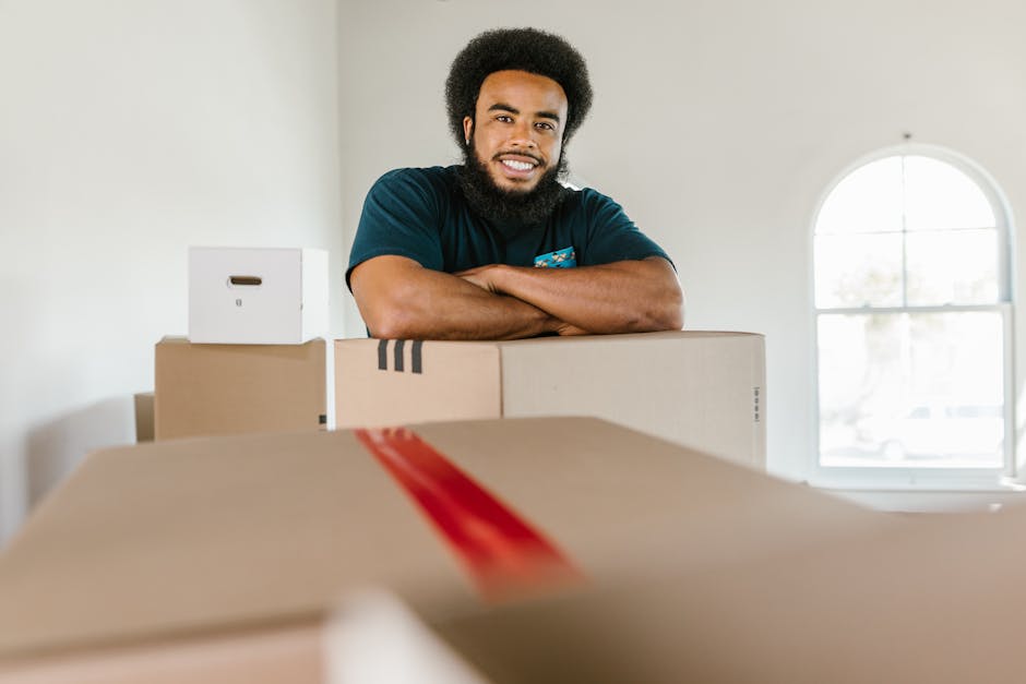 A smiling man with a dark beard and curly hair stands inside a room during a home relocation, resting his arms on top of cardboard boxes of varying sizes, some sealed with red tape. The boxes are arranged on a table or flat surface in front of him, and he appears to be preparing for packing or unpacking. Behind him, the room features a white wall and a large, arched window allowing natural light to illuminate the space. In the background, a white storage box with a handle is visible on a shelf or countertop. The scene depicts the process of furniture transport and packing as part of a removals service, with the environment suggesting an organized moving process facilitated by Man With a Van Harringay, aligning with the context of house removals and moving logistics.