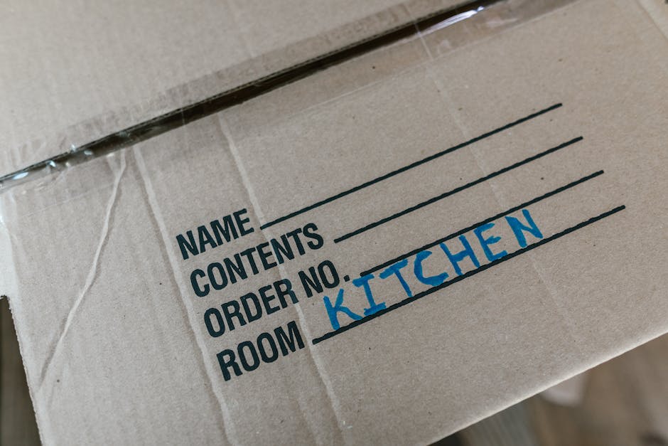 Close-up image of a cardboard moving box with a white label that has black text sections for 'Name', 'Contents', 'Order No.', and 'Room', with 'KITCHEN' handwritten in blue marker in the room section. The box is partially covered with clear packing tape, and the background shows a wooden floor. This box is prepared for home relocation or furniture transport, likely being loaded or transported during a house removal process managed by Man With a Van Harringay, a professional removals service operating in the Haringey area. The image emphasizes packing and moving logistics, with the box situated indoors on a clean floor, indicating careful packing and handling for efficient relocation.
