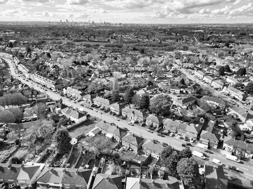 Black and white aerial photograph of a residential neighbourhood showing rows of terraced houses and semi-detached homes along a central street, with cars parked on both sides. The area is bordered by trees, gardens, and backyards, some with visible fencing, sheds, and outdoor furniture. Further in the background, an expansive view of the city skyline with high-rise buildings and a cloudy sky can be seen. The scene captures a quiet suburban environment during daytime, illustrating a typical home relocation setting where houses are prepared for packing and furniture transport. The image highlights the dense arrangement of dwellings and the open spaces that facilitate the loading process of a house removal service, such as those offered by Man With a Van Harringay, as part of their comprehensive removals and packing solutions.