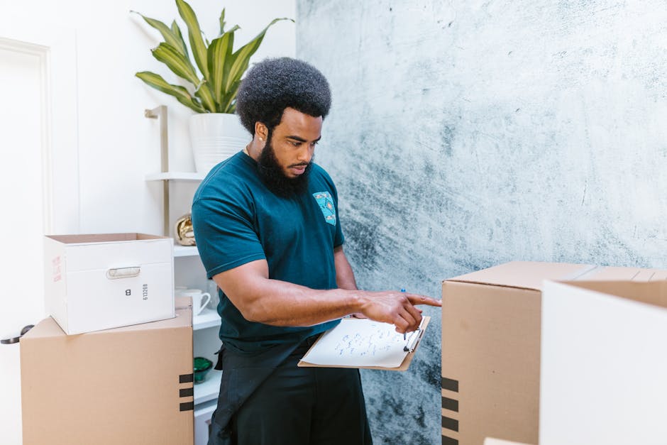 A man with a dark curly hair and beard, dressed in a navy blue t-shirt, is inside a room, holding a clipboard in his left hand and pointing with his right index finger towards a cardboard box. The room contains several packed cardboard boxes, some sealed with packing tape, and a white shelving unit with a potted plant placed on top. The background wall has a textured light blue and white finish, and the lighting is natural and evenly distributed. The man appears to be reviewing a packing or loading checklist as part of a home relocation or furniture transport process, with the boxes arranged around him for moving or storage purposes. This setting reflects aspects of professional removals, including packing, organization, and the coordination involved in loading items for transport, consistent with services provided by Man With a Van Harringay during house removals or moving logistics.