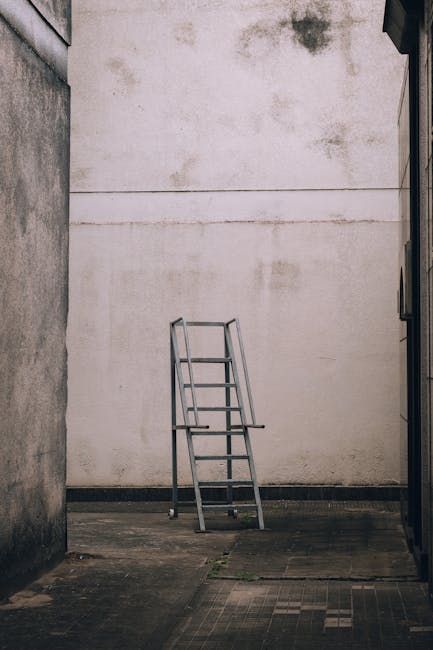 A metal ladder with four steps positioned outdoors against a plain, weathered white wall in a narrow alleyway. The ground is concrete with some visible dirt and minor cracks. The alley is flanked by two tall building walls, one on the left with a rough texture and the other possibly a smooth brick or concrete surface partially visible on the right side. The lighting is natural, suggesting daytime, and there are no other objects or people in the scene. This image might relate to home relocation or packing and moving activities, illustrating a secure method for accessing higher areas, relevant to furniture transport and removal planning, as presented by Man With a Van Harringay.