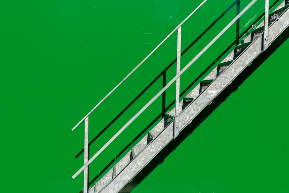 Aerial view of a metal staircase with black non-slip treads and silver handrails, positioned against a bright green outdoor surface. The staircase is used for access between levels of a building involved in a home relocation process, with the surrounding area indicating an outdoor environment. The stairs are being prepared for moving or loading activities, related to furniture transport and packing and moving services. The photograph shows the steps descending from an upper level to a lower paved area, with sunlight casting shadows on the metal structure, emphasizing the staircase's construction details. The image is associated with professional removals performed by Man With a Van Harringay, highlighting the logistical aspects of furniture and box transport during house removals involving tight staircases or limited access points.