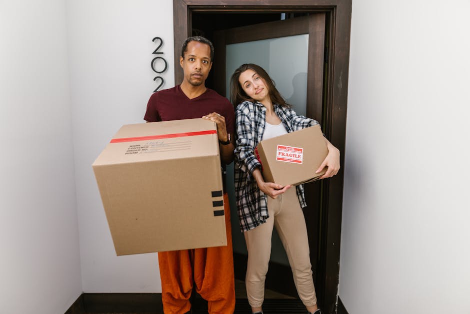 A man and a woman standing inside a residential doorway during a home relocation process. The man, dressed in a maroon shirt and orange trousers, is holding a large cardboard box with packing tape and markings, indicating it is part of the packing and moving process. The woman, wearing a plaid shirt over a white top and beige trousers, is holding a smaller cardboard box labeled with a 'Fragile' sticker. Both individuals are positioned on a narrow interior corridor with white walls and a dark wooden doorframe. The lighting appears natural and even, highlighting the packing materials, including cardboard boxes of various sizes, which are part of the furniture transport and packing process. The setting reflects activities associated with removals services, such as loading or unloading boxes during a house move, supporting the focus of Man With a Van Harringay in house removals and parking permit tips for the N4 postcode area.