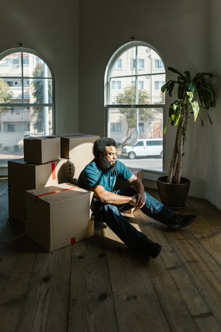 A man with dark curly hair, wearing a blue t-shirt and dark trousers, sitting on a wooden floor inside a room with large arched windows that let in natural daylight. He appears to be resting amid several cardboard boxes that are sealed with red packing tape, some of which are stacked behind him. The room has a tall potted plant placed near one of the windows, contributing to a minimal yet functional interior setting typical of a home or an office preparing for a domestic move. Outside the windows, a view of nearby buildings and a parked white vehicle is visible. This scene may be part of a home relocation process, illustrating the stage during packing or the transition area before loading furniture and boxes into a vehicle for transport, aligning with the services provided by Man With a Van Harringay in house removals and furniture transport.